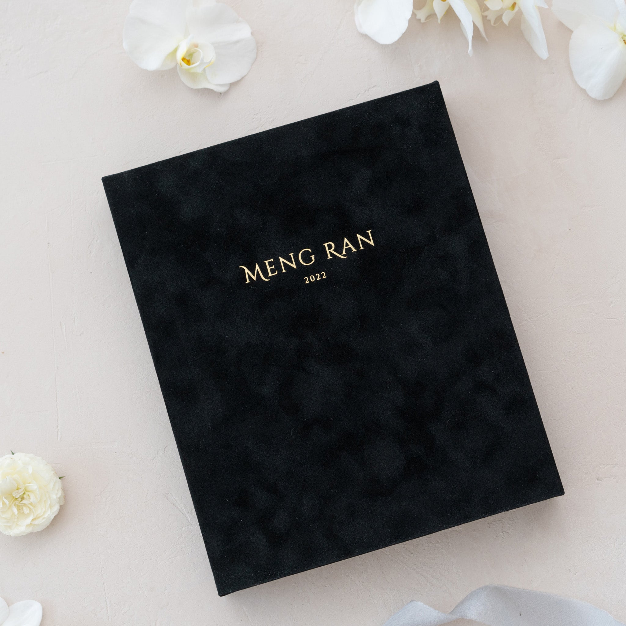 A black photo album with gold text on the cover, placed on a surface with white flowers.