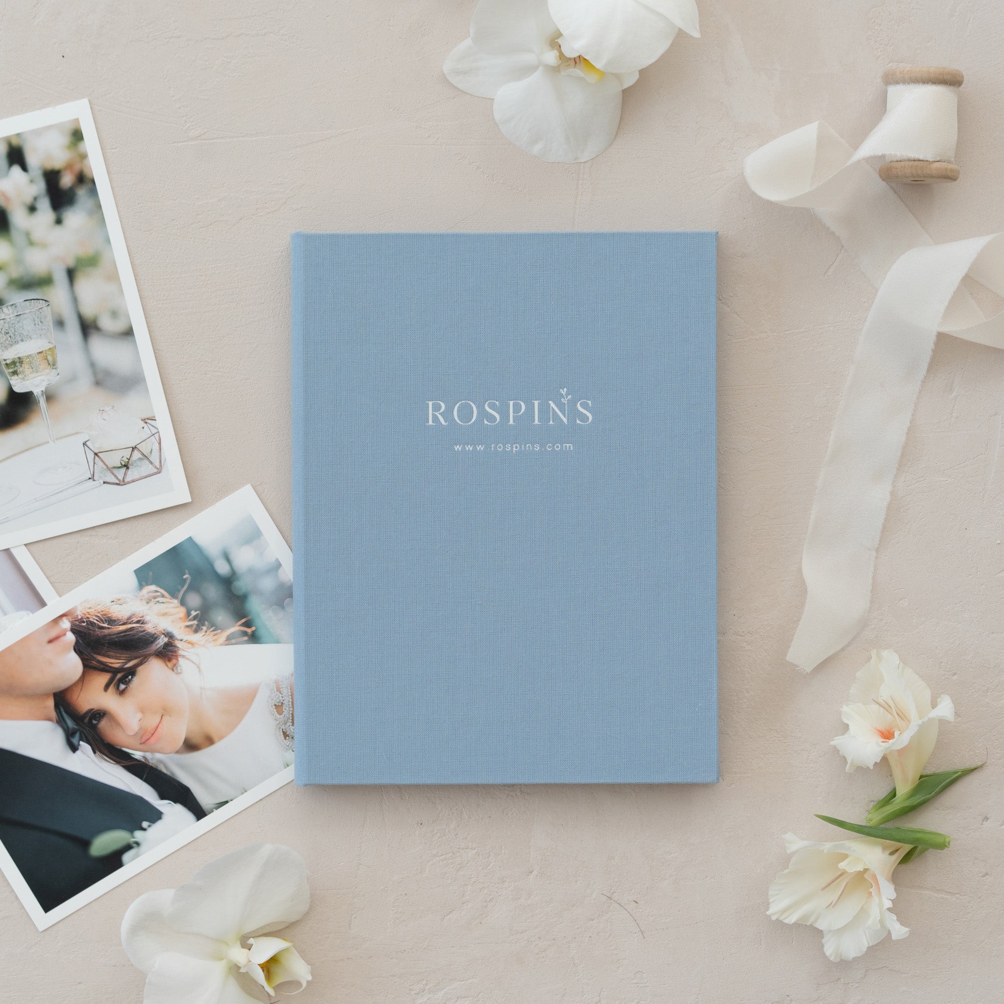 a blue book with the text "ROSPIN'S" on it, surrounded by various items such as photographs, flowers, and a ribbon.
