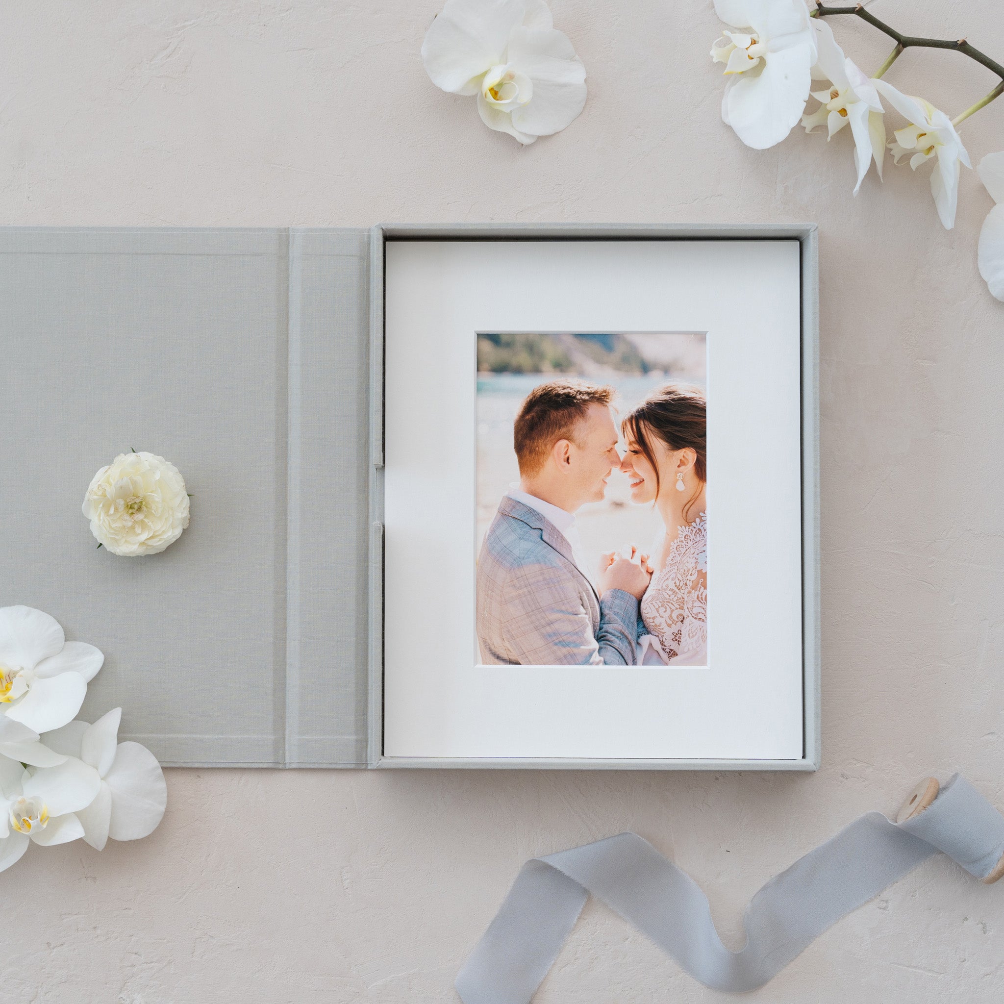a gray photo album with a picture of a couple inside, surrounded by white flowers and a ribbon.