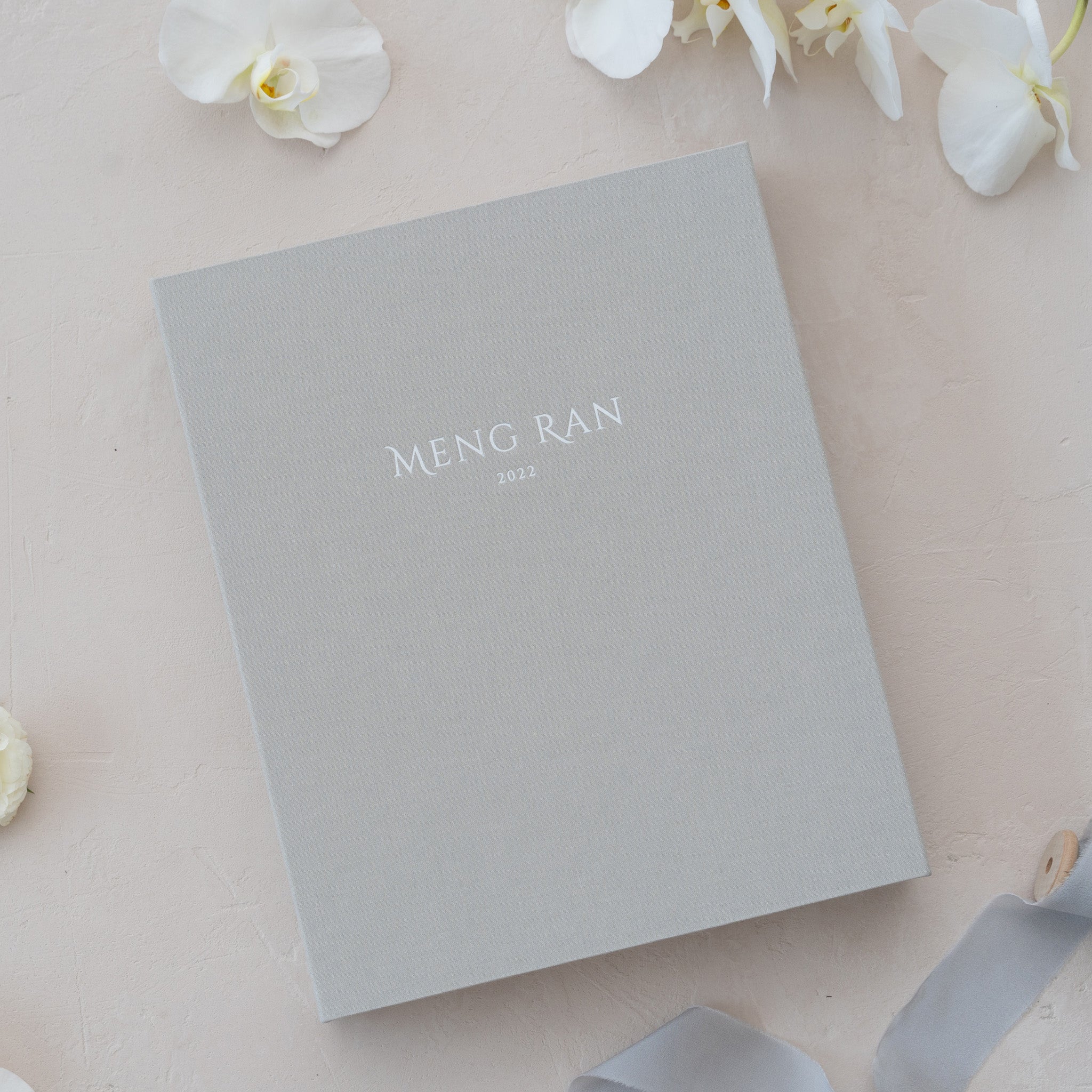 A gray photo album with the name 'Meng Ran' printed on it, placed on a light-colored surface with white flowers in the background.