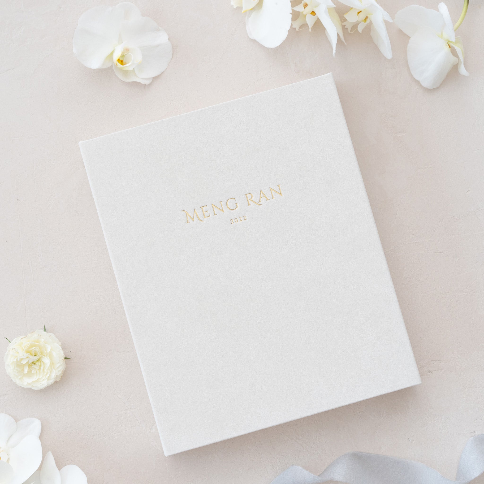 A white photo album with the text "Meng Ran" printed on it, surrounded by white flowers on a light-colored background.
