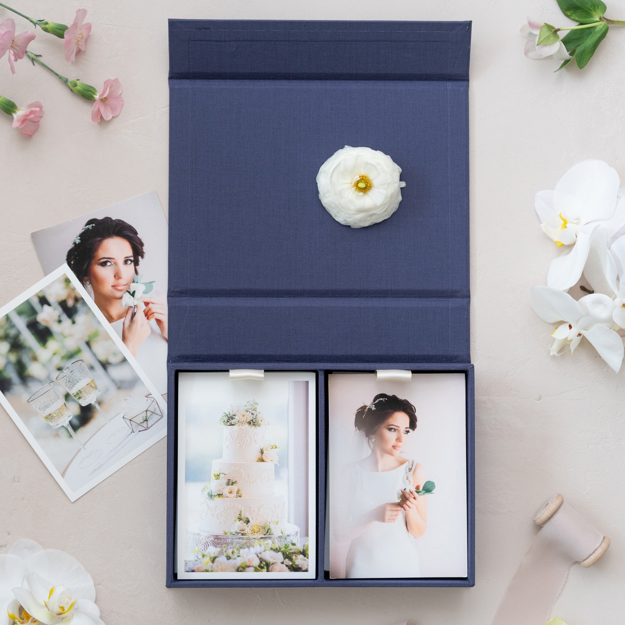 a blue photo album with two photographs of a woman, one of which is a wedding cake. The album is surrounded by various flowers, including white and pink flowers, and a single white flower.