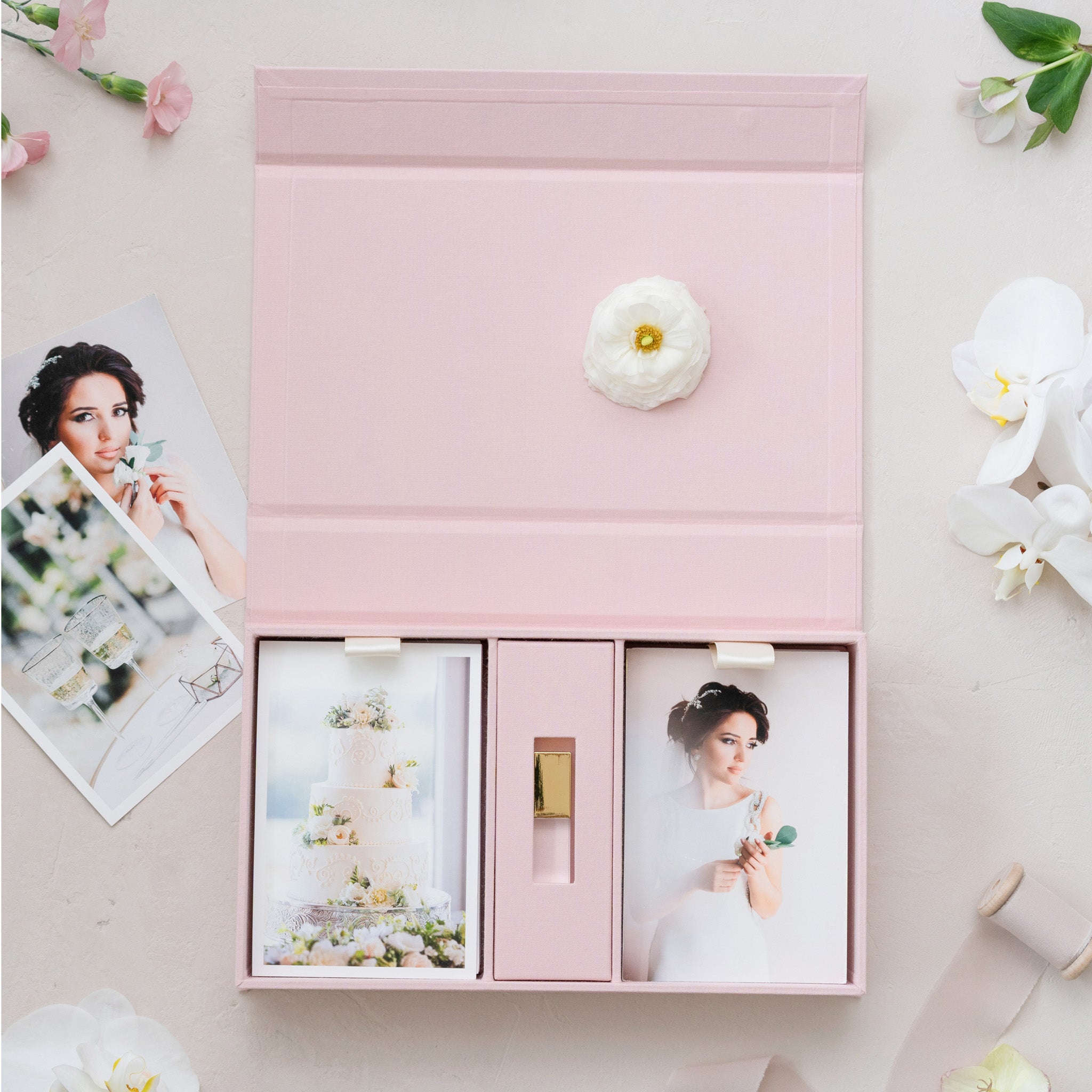 a pink box containing two photographs of a woman, one of which is a wedding cake. The box is surrounded by various flowers, including white orchids, and there are also some photographs scattered around the box.