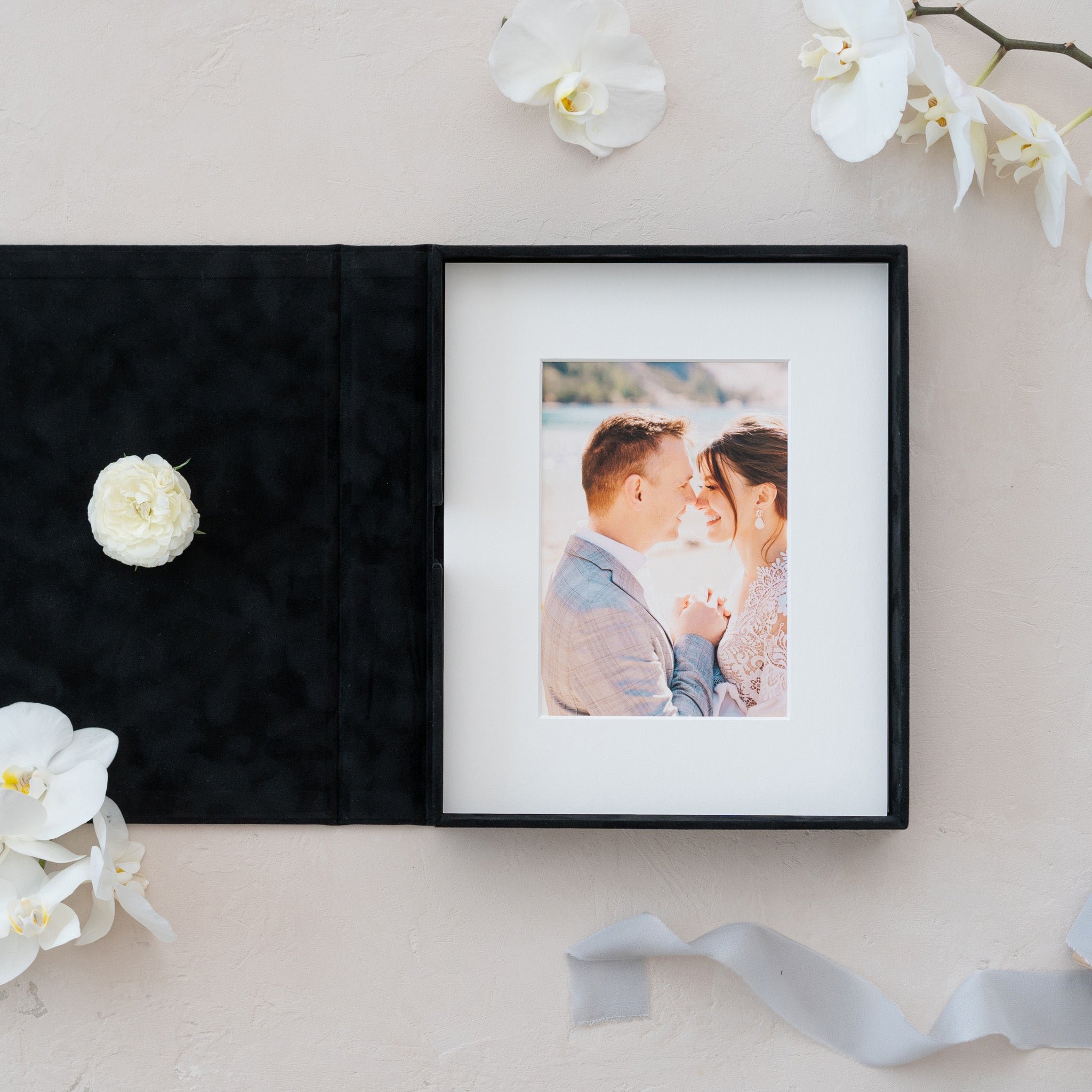 a black photo album with a framed photograph of a couple, placed on a white surface. There are also white flowers and a ribbon nearby, creating a romantic and elegant atmosphere.
