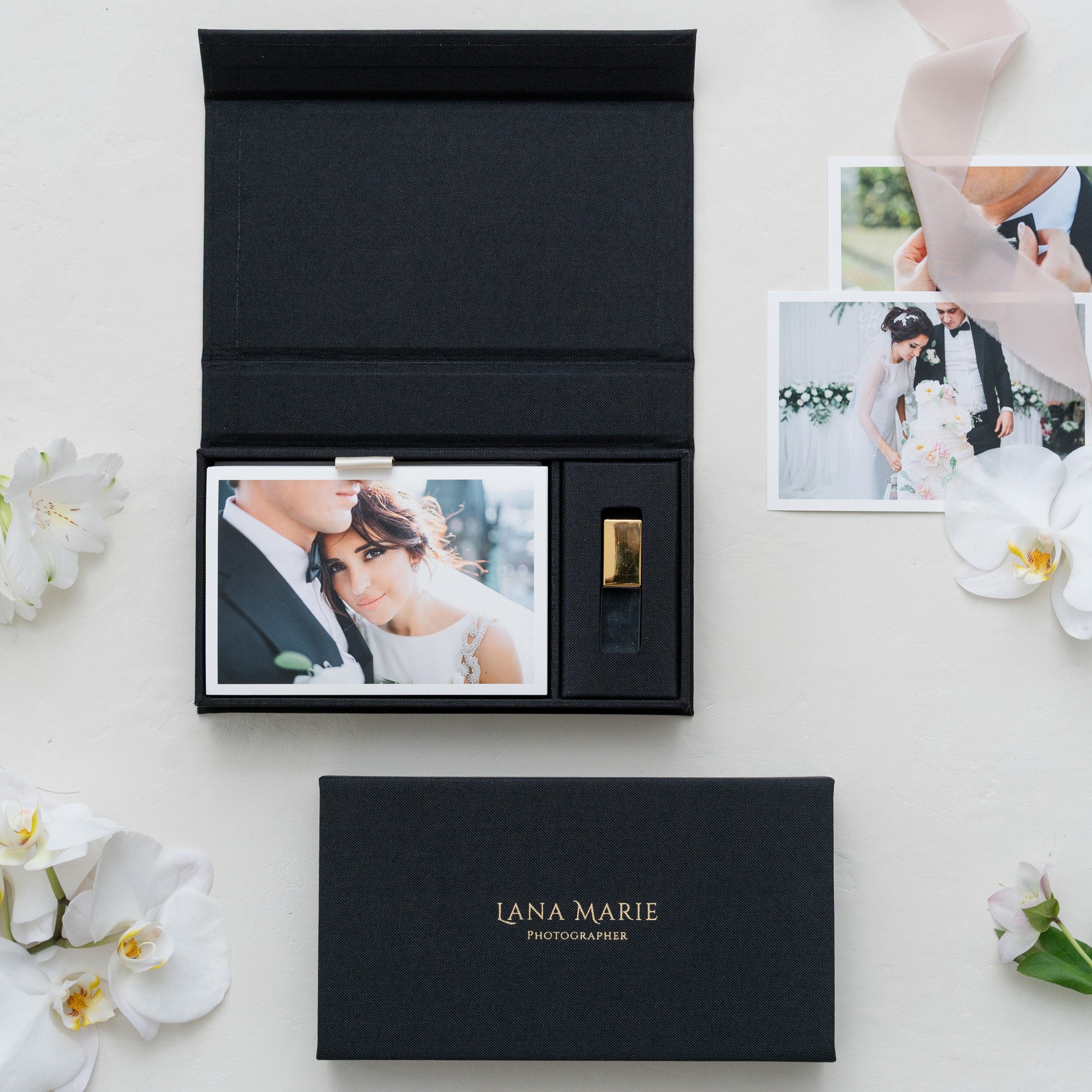 a black photo box with a gold-colored USB flash drive inside, surrounded by white flowers and a few photographs.