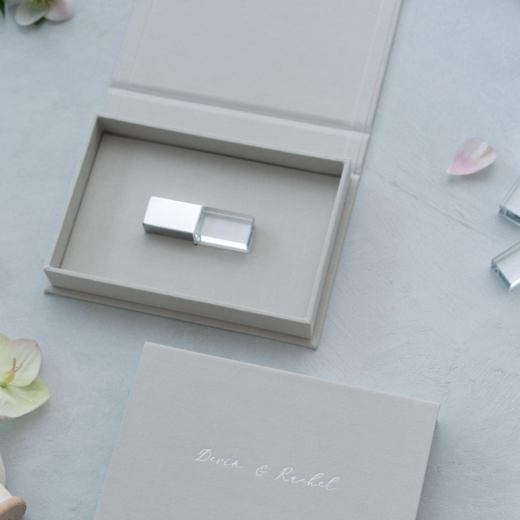 a USB flash drive placed inside a grey box, with a small pink flower and some other objects on a light blue surface.