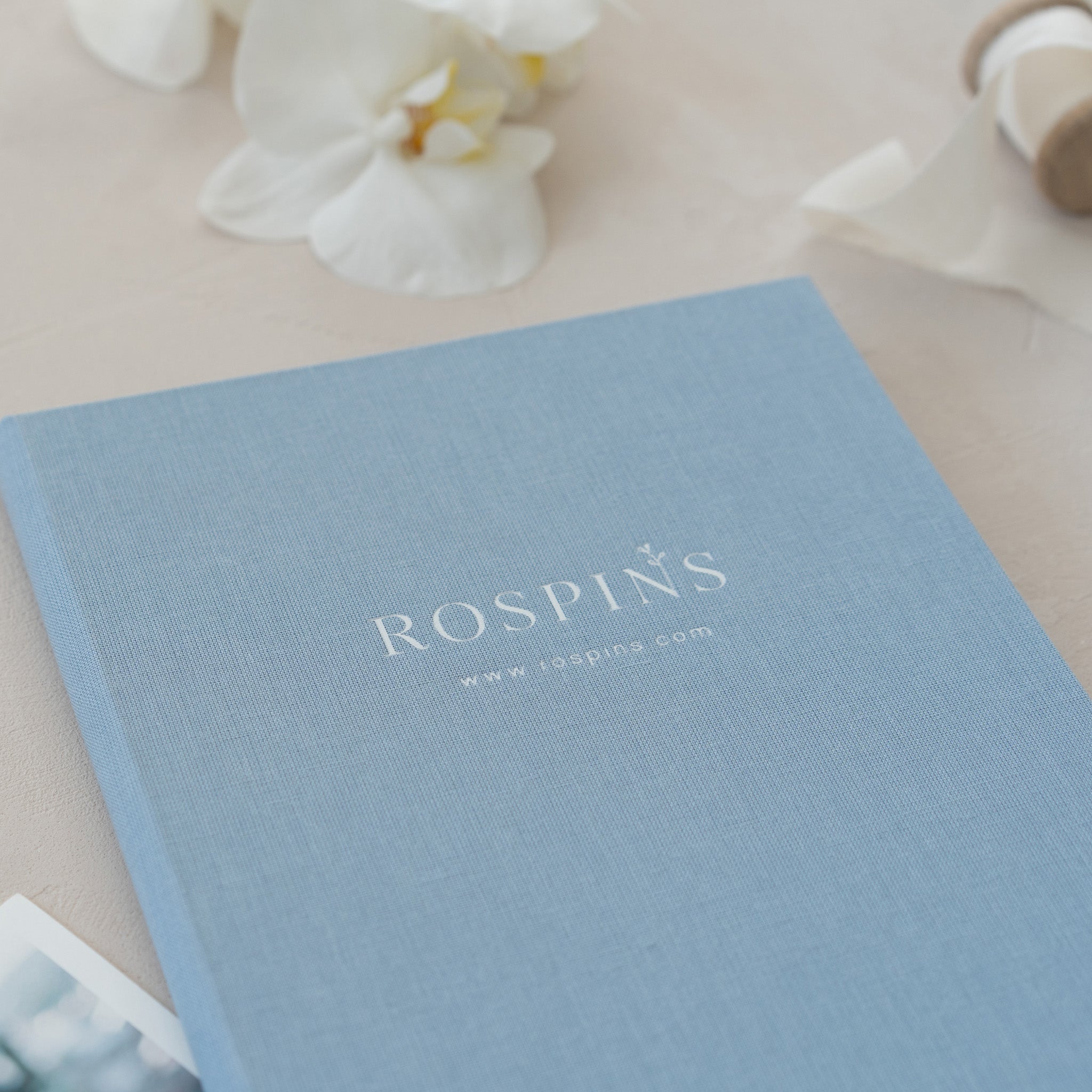 A light blue book titled "ROSPIN'S" with a website address "www.rospin.com" printed on it, placed on a table with white flowers in the background.