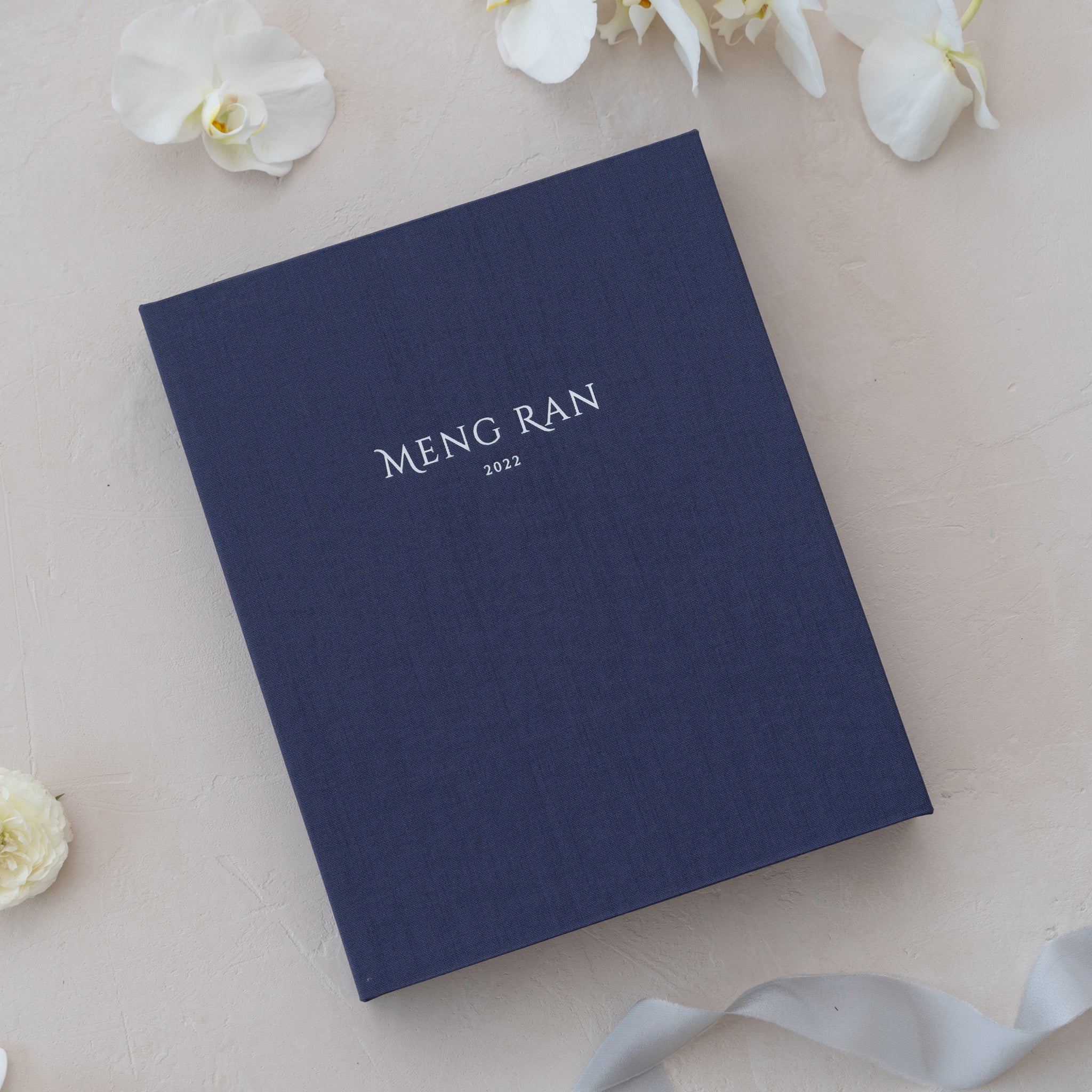A navy blue photo album with the title "Meng Ran 2012" printed on the cover, placed on a surface with white orchid petals and a ribbon.