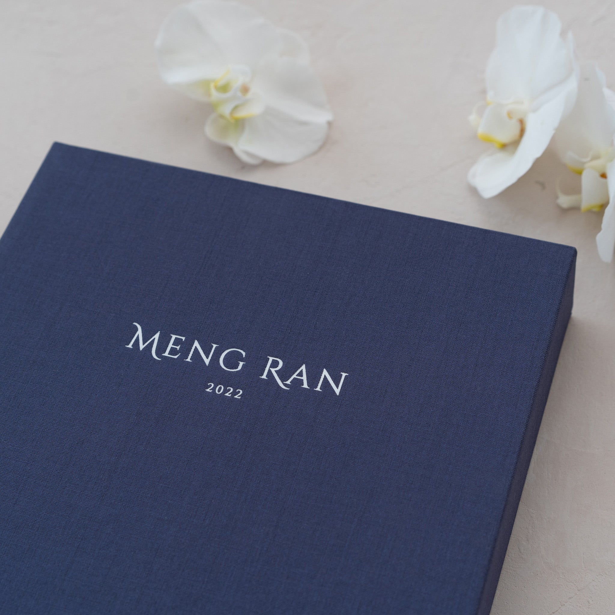 A navy blue box with the name 'Meng Ran' and the year '2022' printed on it, along with a few white orchid flowers.