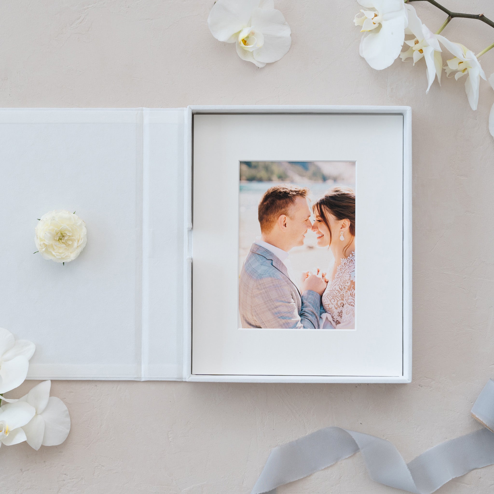 a white photo frame containing a photograph of a couple, placed on a light-colored surface. Surrounding the frame are white flowers, and a ribbon is visible in the bottom right corner of the frame.
