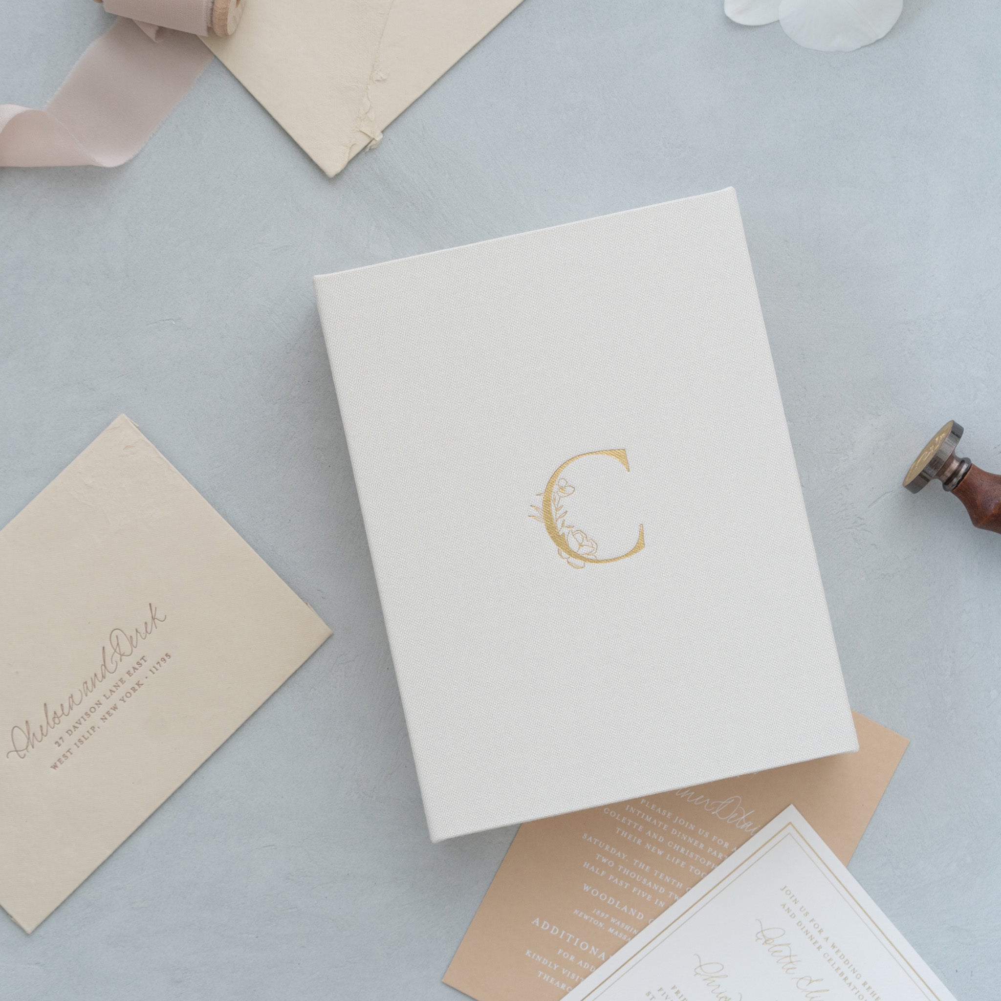various stationery items, including a white envelope with a gold letter "C" on it, a white card, and some other papers, all placed on a light blue background.